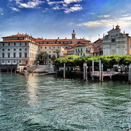 Historic buildings with red roofs and a white church by a waterfront with several docks, under a partly cloudy sky.