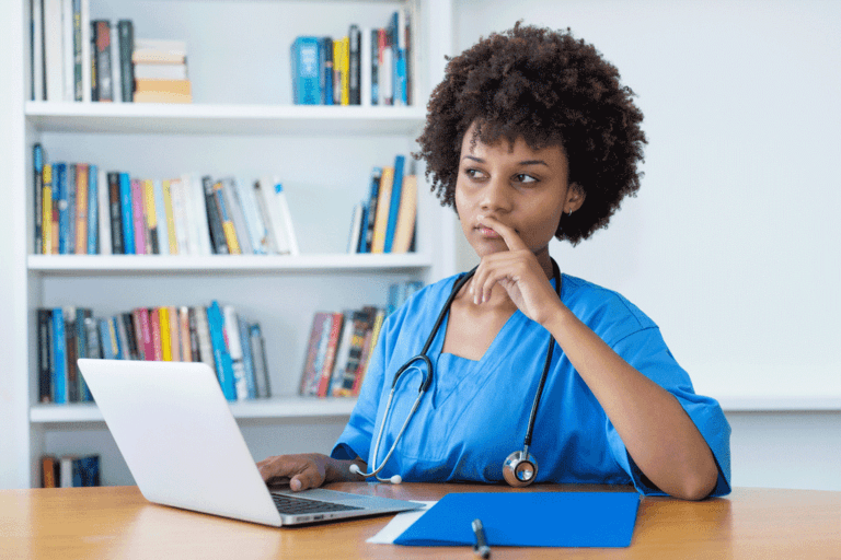 Nurse in blue scrubs sitting at a desk with a laptop, thinking with hand on chin.