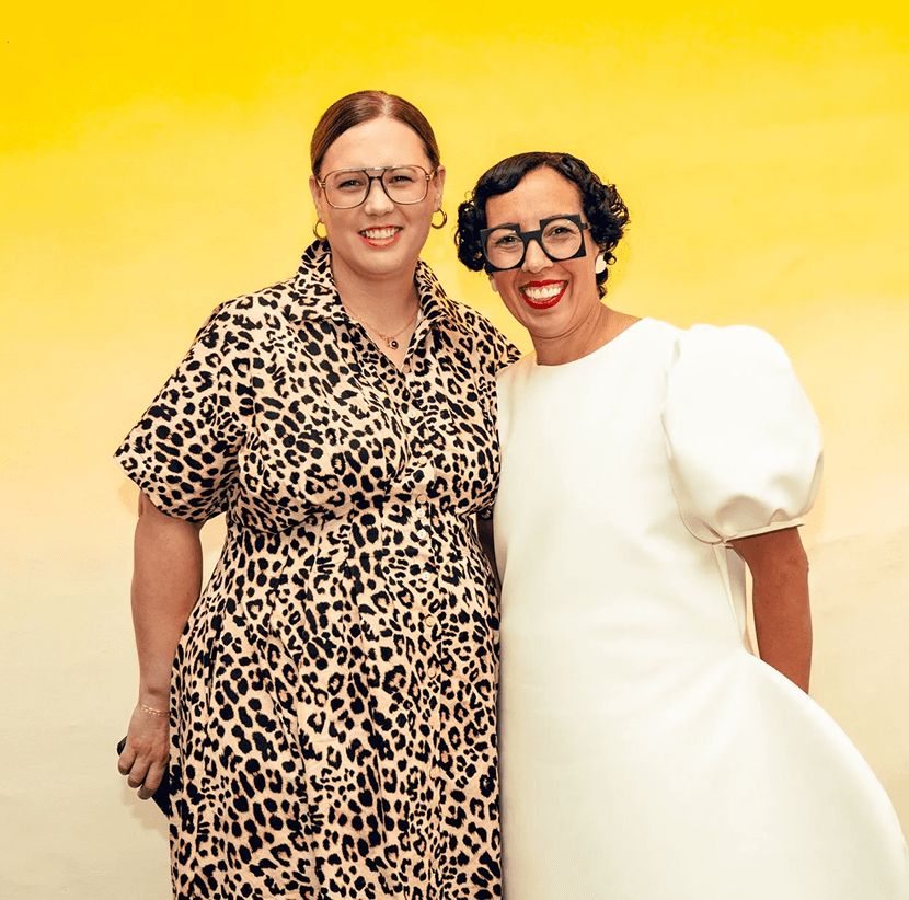 Two women stand together, smiling, against a bright yellow background. One wears a patterned dress, the other a white outfit.