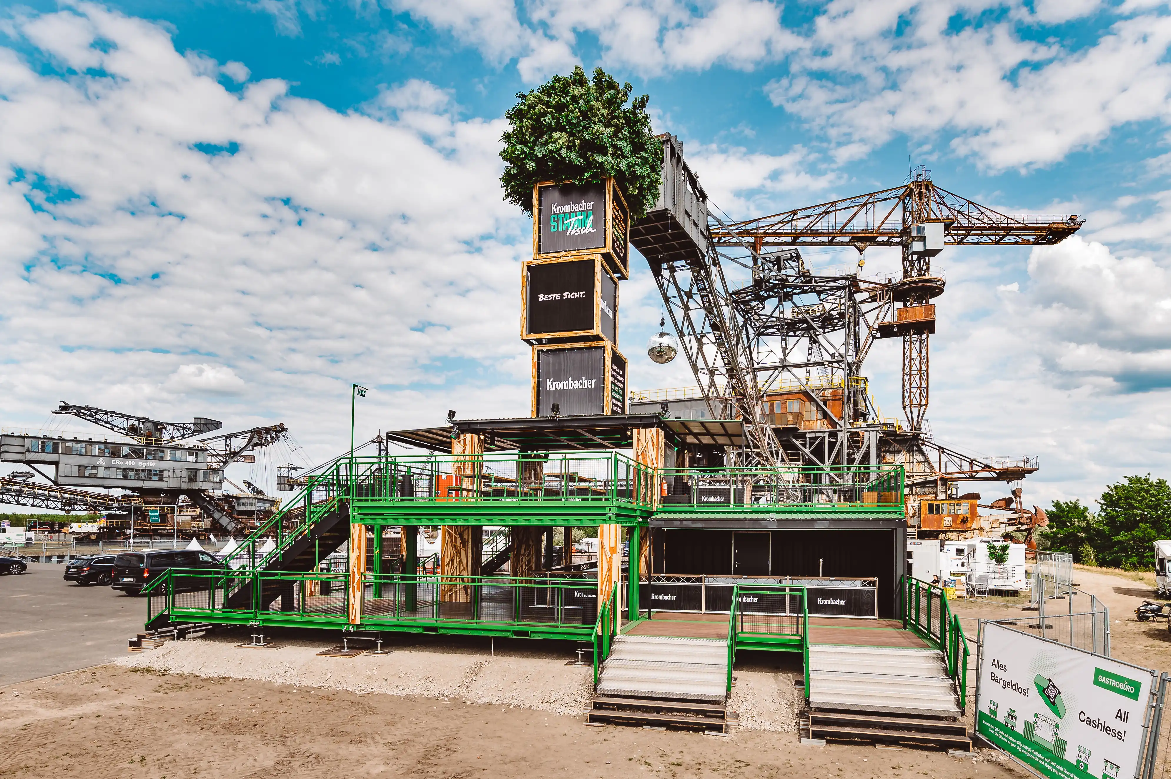 Wide view of the KROMBACHER stand at Ferropolis with crate tower, green scaffold terraces and industrial cranes under a blue summer sky.