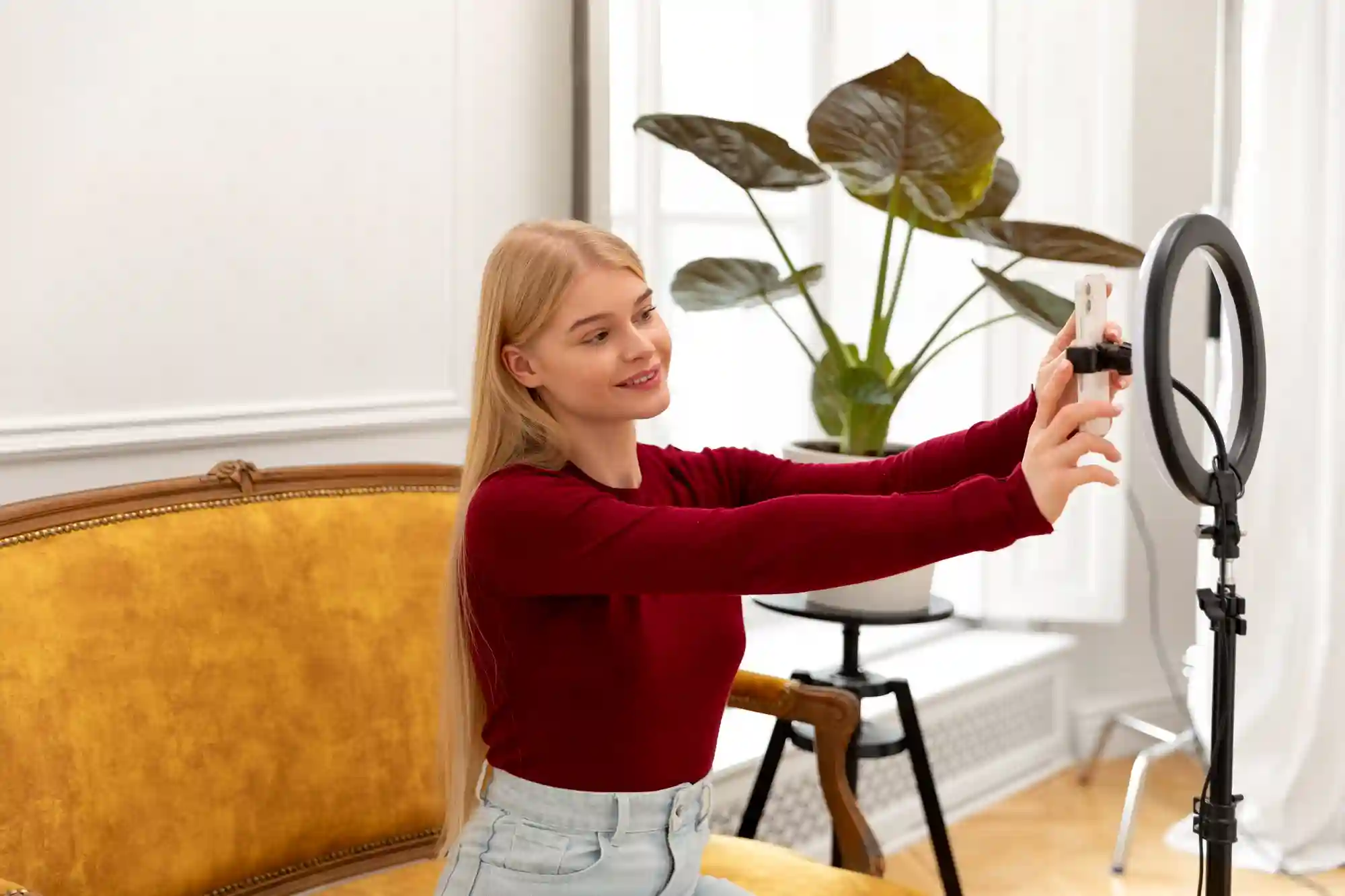 Young blonde woman recording video content with a smartphone mounted on a ring light indoors.