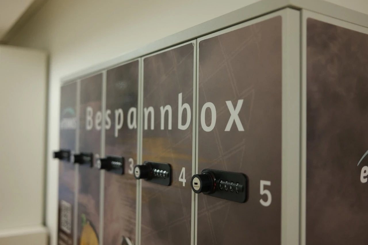 Eine Bespannbox mit fünf nummerierten Fächern, die jeweils mit einem Zahlenschloss gesichert sind. Die Box ist mit einem Tennis-Motiv bedruckt und trägt das Branding von “eTENNIS” und “Stefs Racket Corner”. Ein QR-Code auf der linken Seite weist darauf hin, dass Kunden ihre Schläger für eine Bespannung abgeben können.