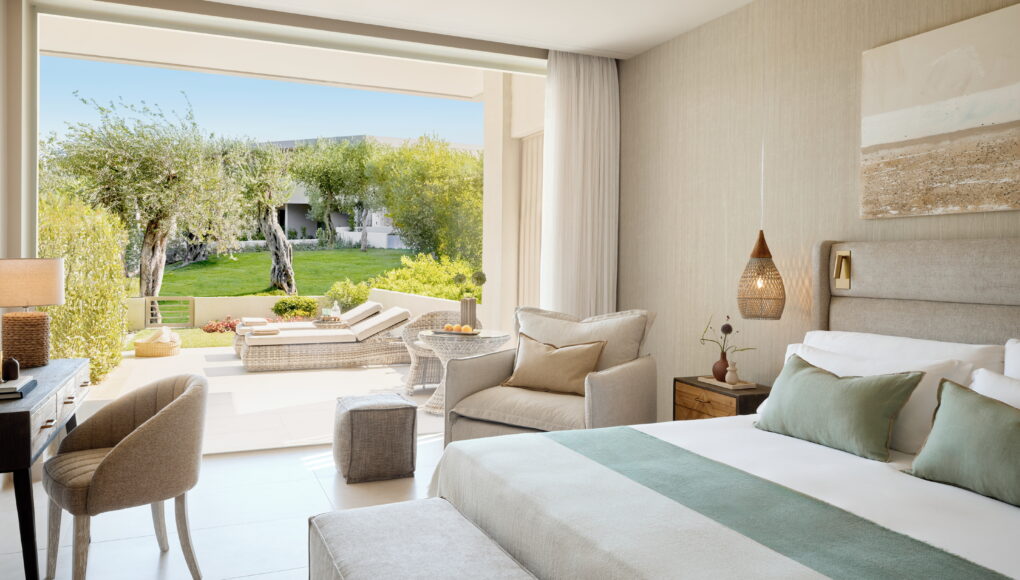 Helles, modernes Hotelzimmer mit großem Doppelbett, gemütlichem Lesesessel und direktem Zugang zu einer Terrasse mit Gartenblick.