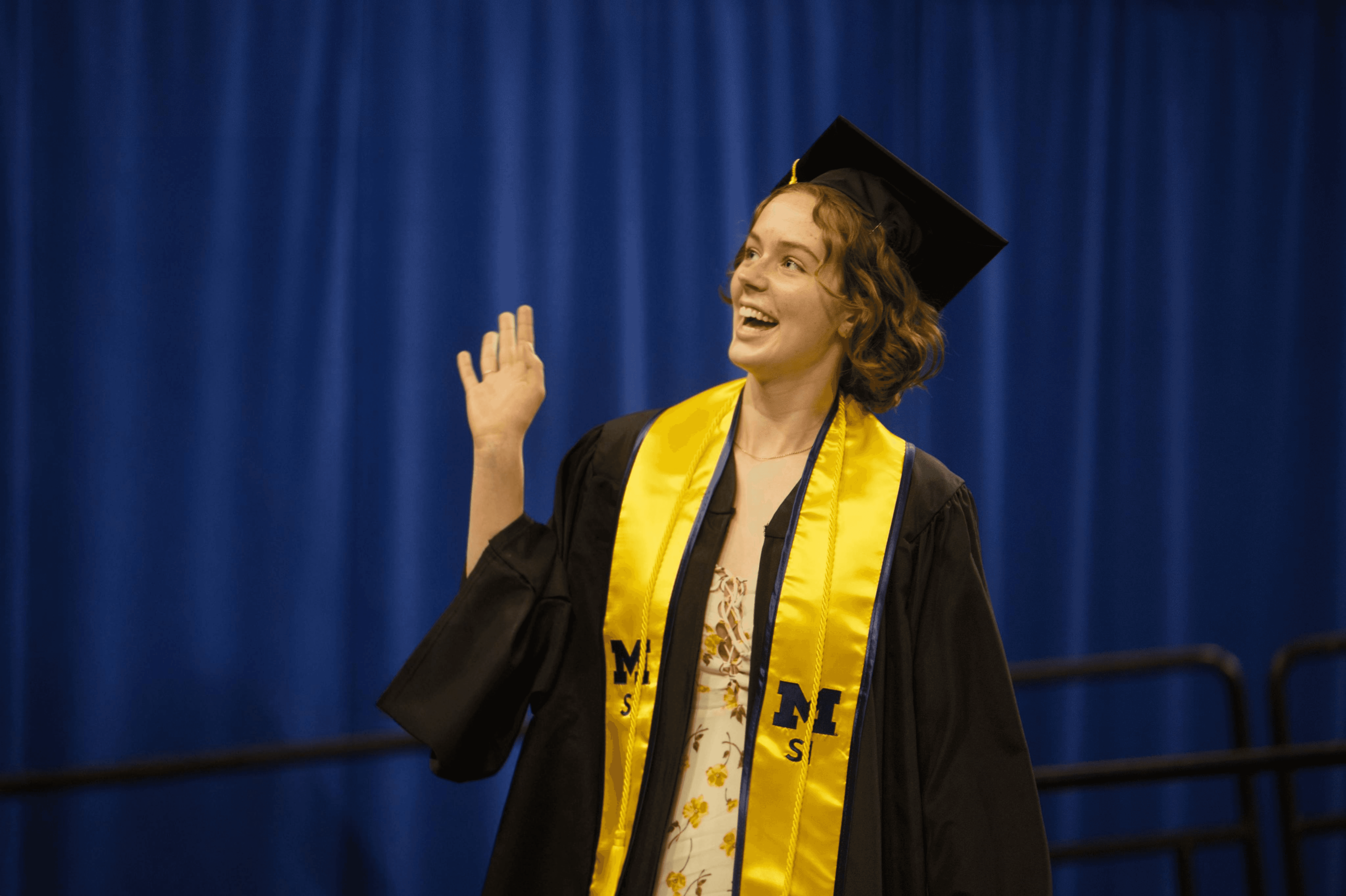 Maddie waving in front of a blue curtain, wearing a graduation cap and gown and University of Michigan stole.