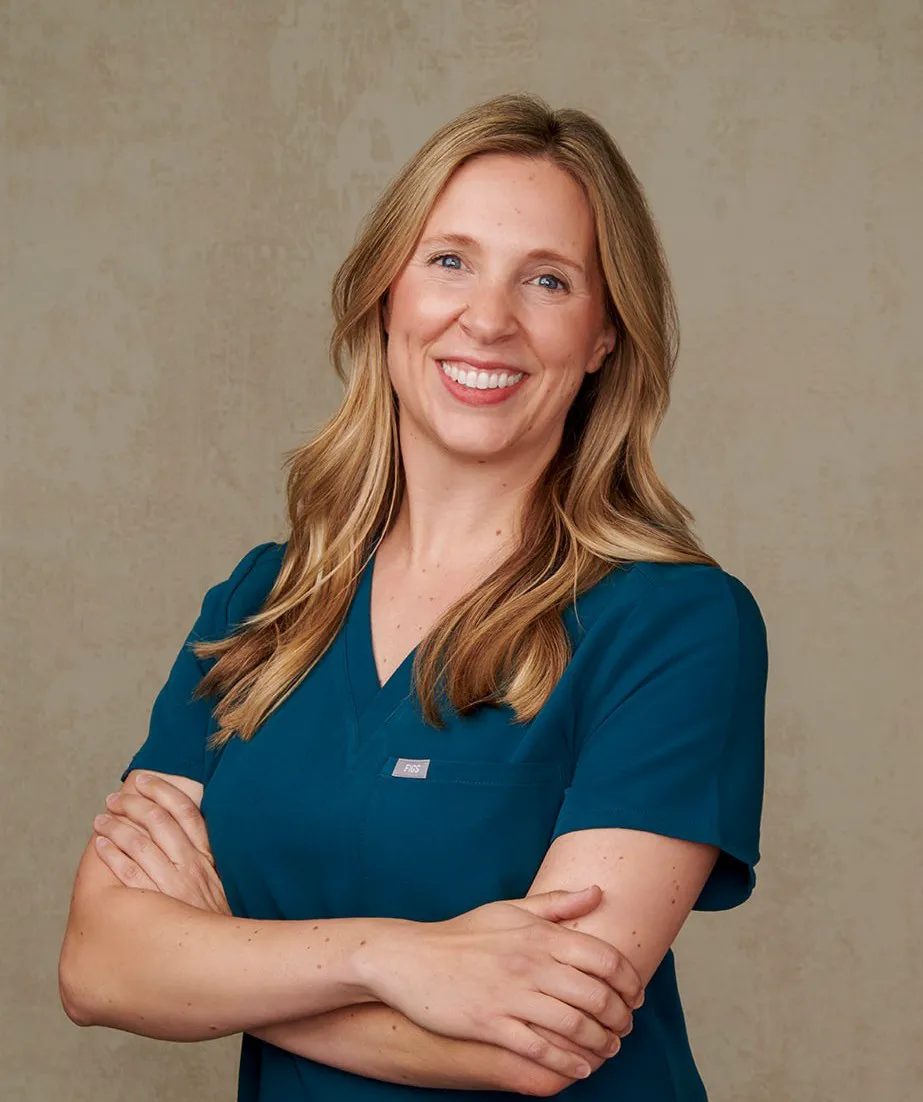 Trish Mueller, founder of Lift and Revive, smiling with arms crossed in teal medical scrubs against a textured beige background