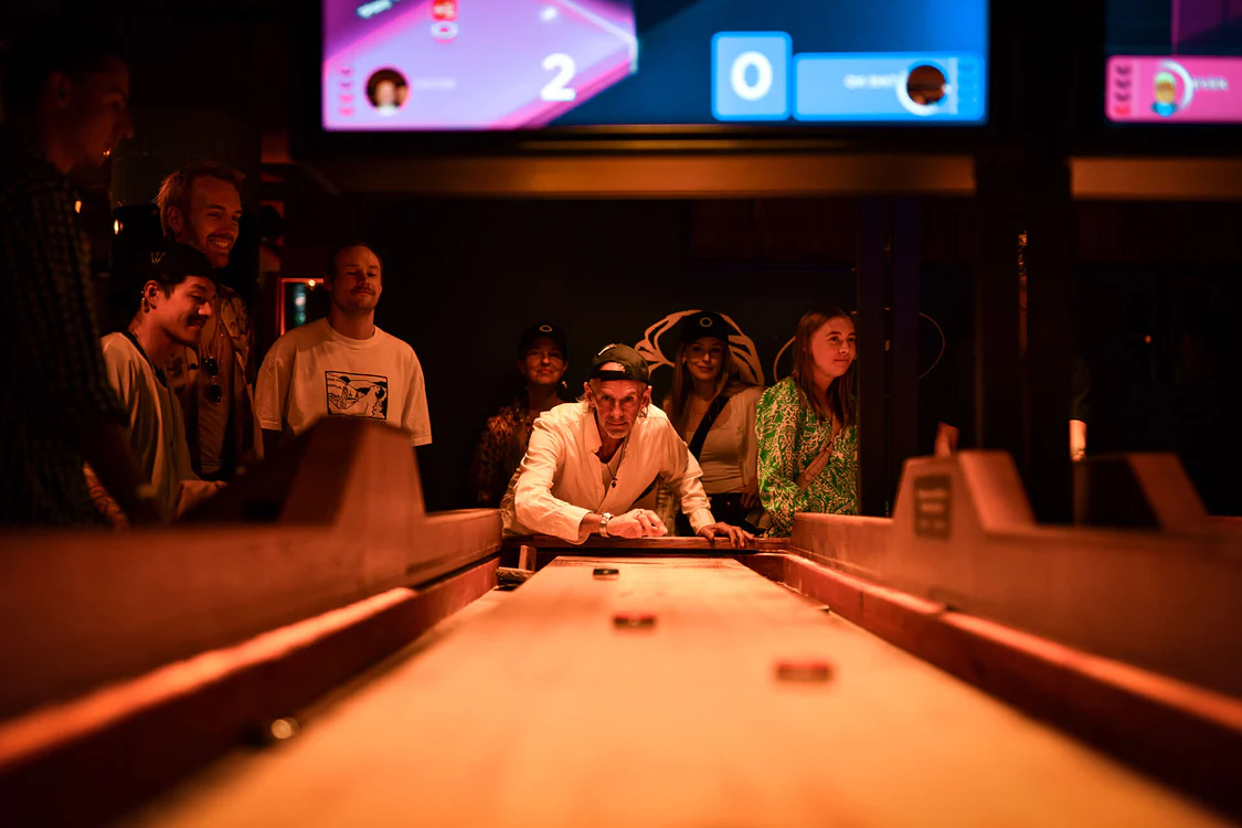 A young person with curly hair smiles at the camera while sitting in front of a laptop against a dark background.