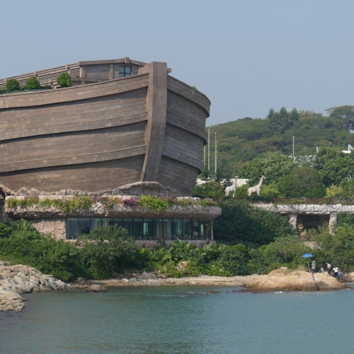 Egy nagy, csónak alakú épület a víz mellett, zöldövezettel és állatszobrokkal a környező területen.