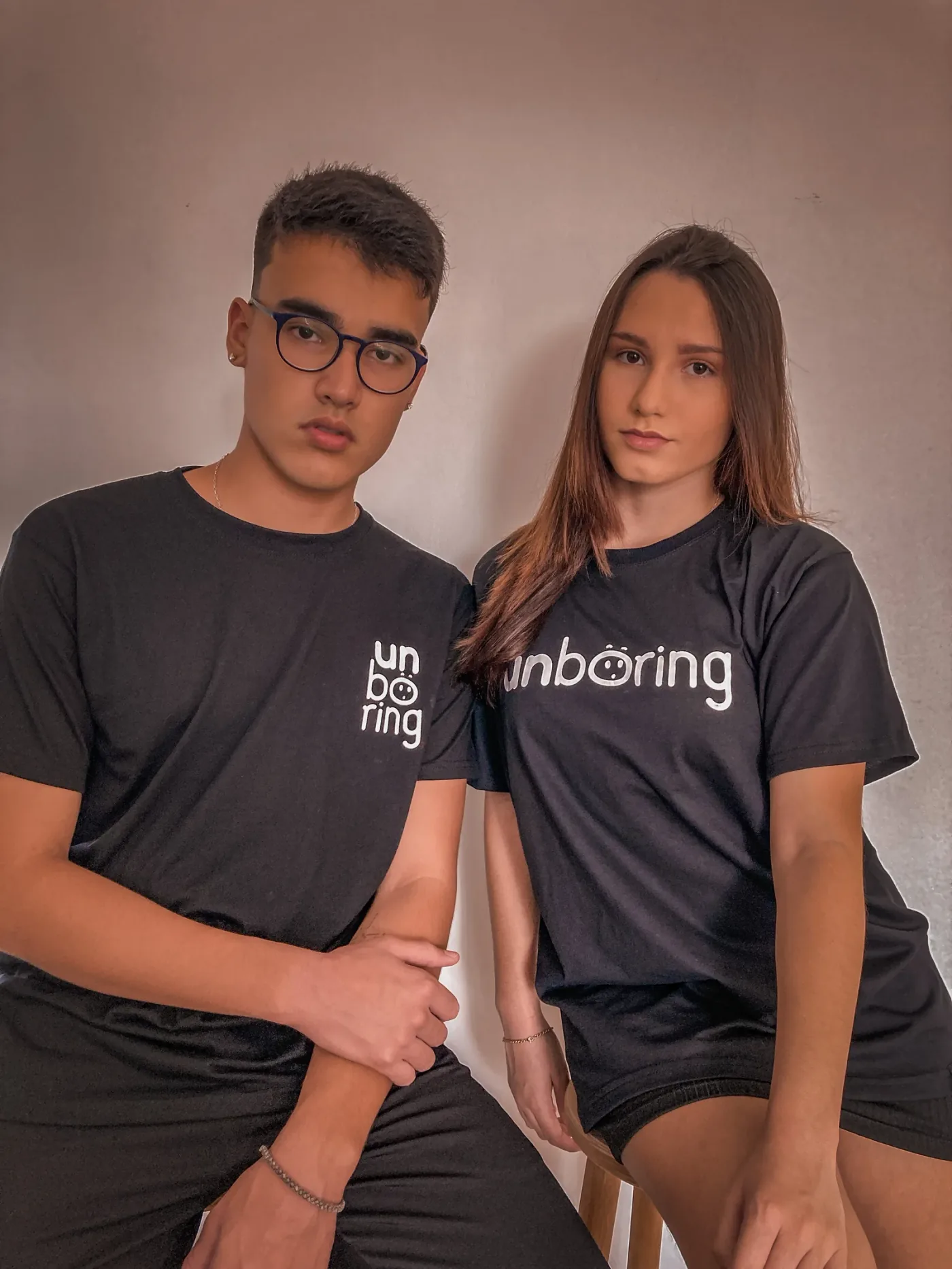 A young man and woman pose against a plain wall in black Unboring T-shirts. His T-shirt uses the stacked wordmark on the left chest; hers carries the horizontal wordmark across the chest.