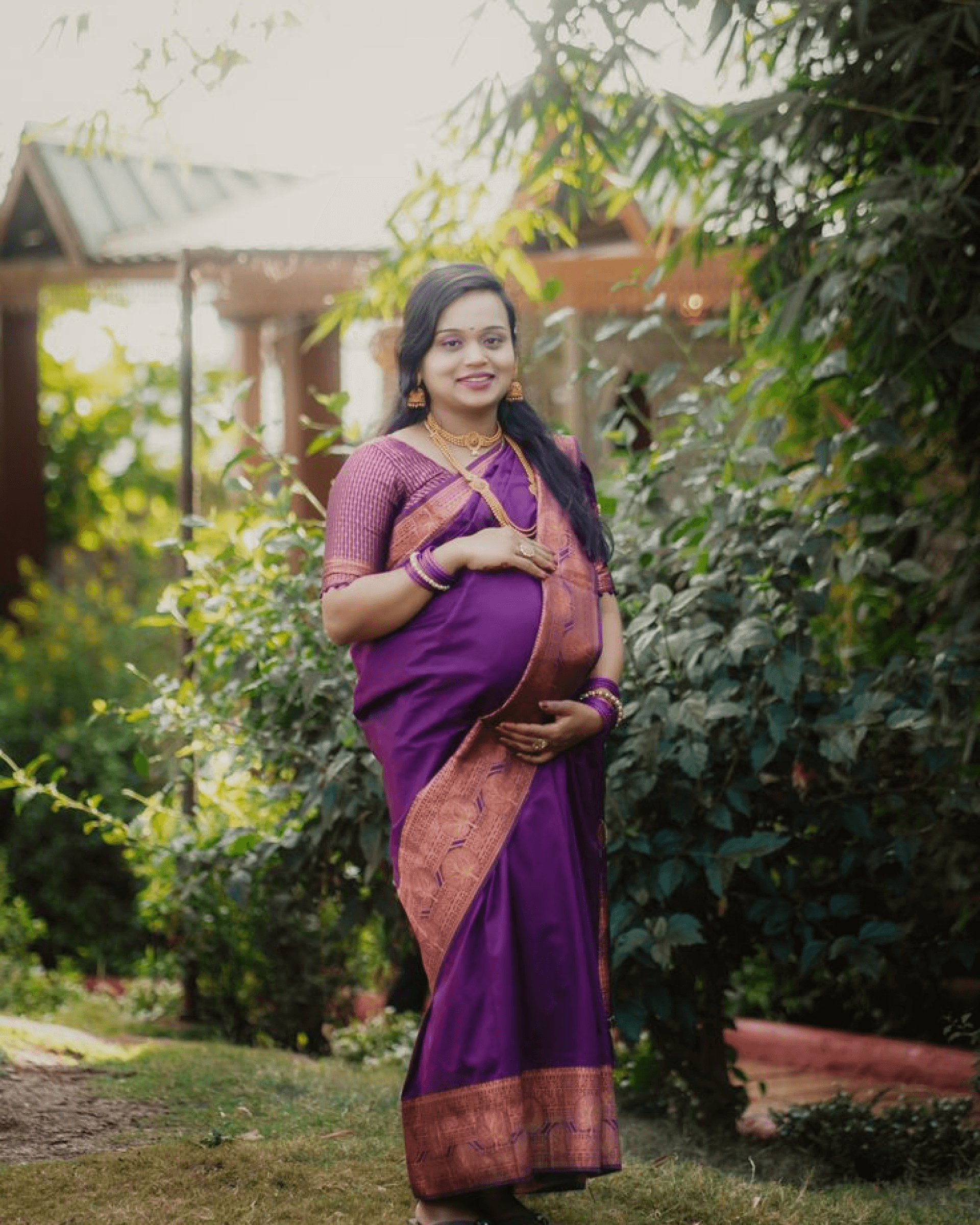 Pregnant woman in a purple saree standing outdoors and smiling confidently.
