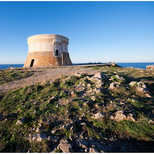 Ett gammalt, cylindriskt stenstorn står på en stenig, gräsbevuxen kulle med en klarblå himmel och havet i bakgrunden.