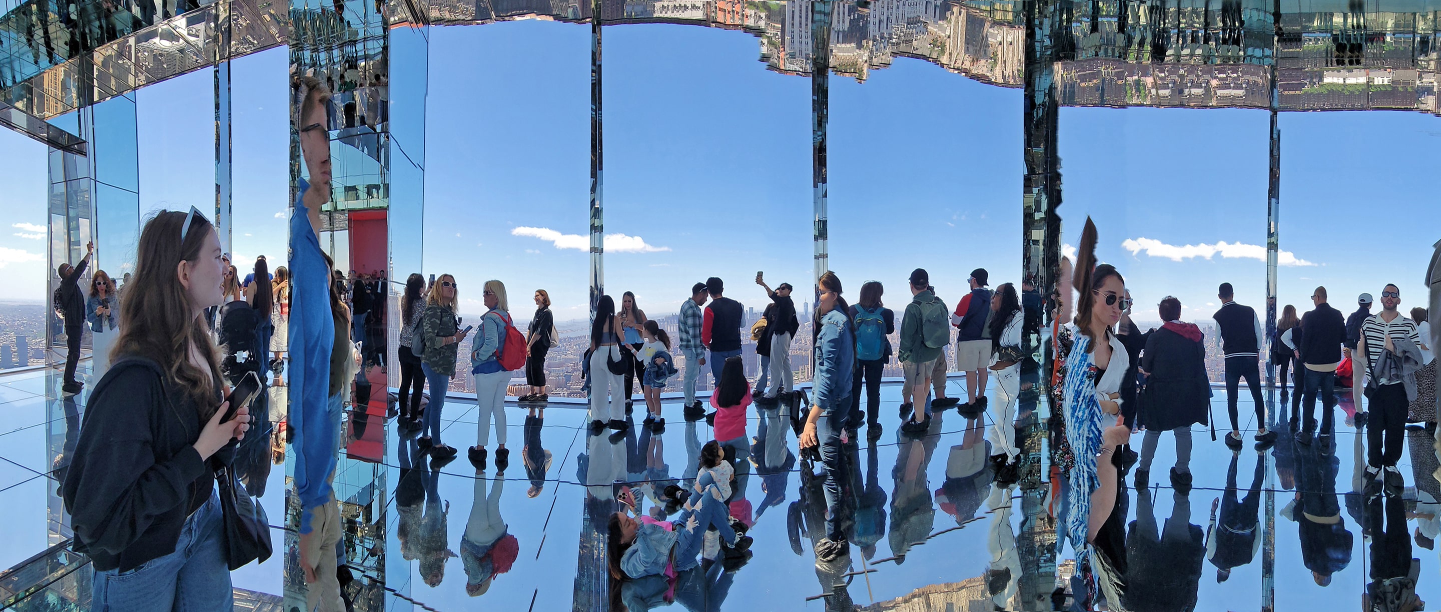 Panoramic photograph inside a mirrored observation deck where visitors are reflected across glass walls and polished floors. The expansive sky outside contrasts with the fragmented reflections, creating a surreal interplay between light, perspective, and presence.