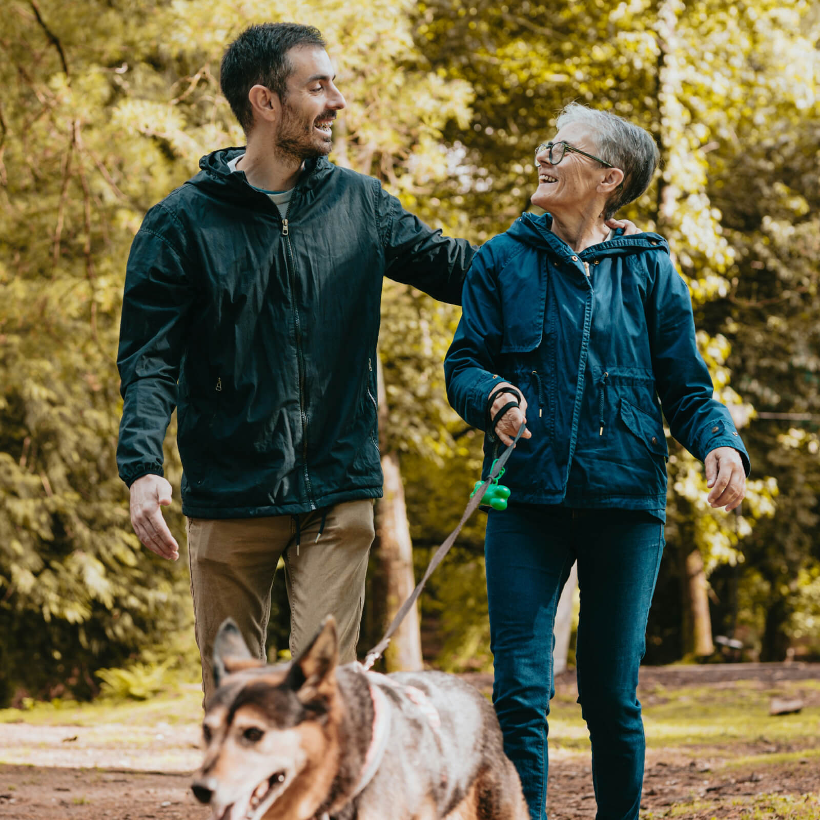 Ein Mann und eine Frau zugewandt zueinenader im Gesprcäh, gehen mit einem Hund an der Leine durch den Wald spazieren