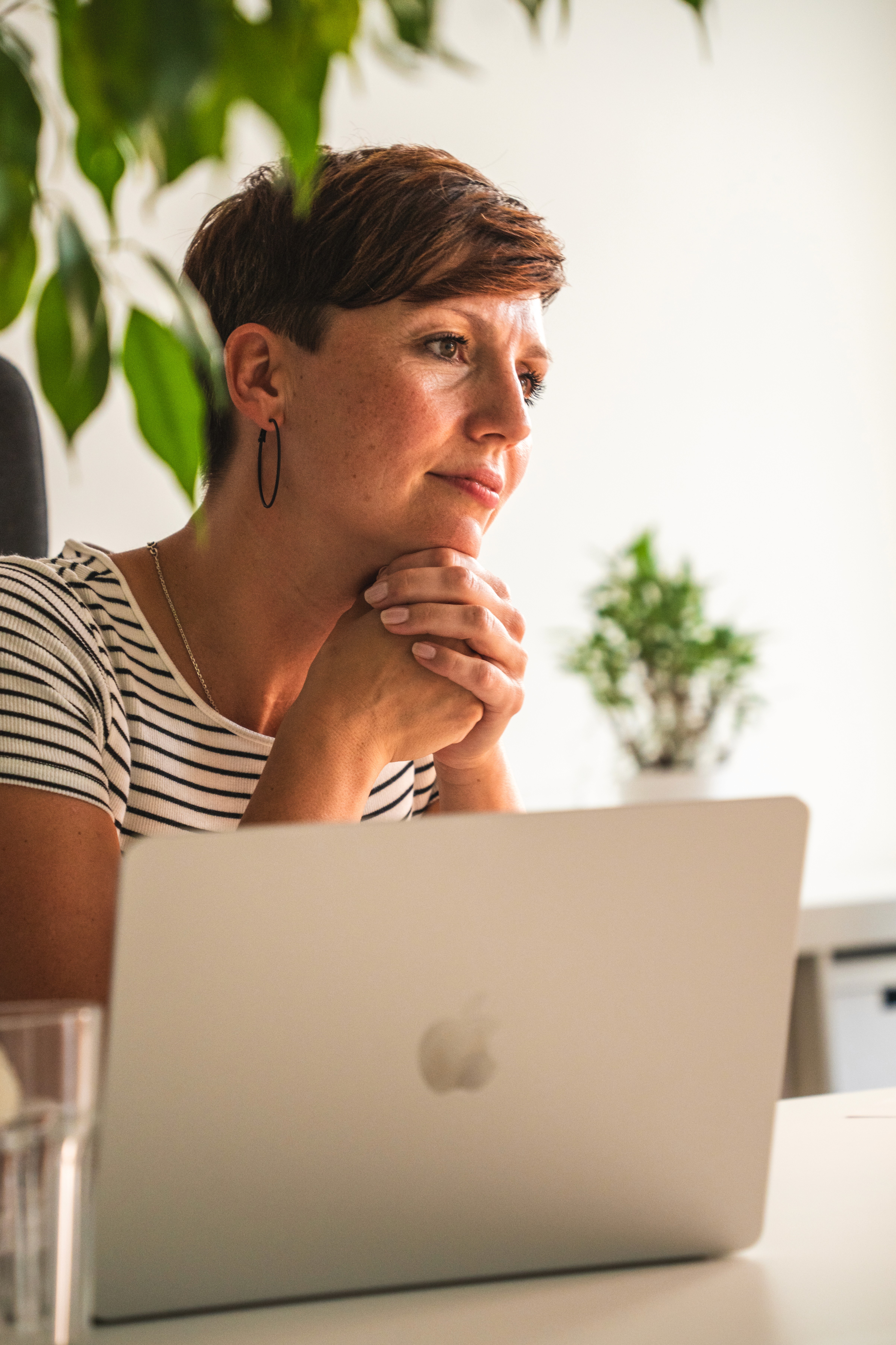 Person mit kurzem Haar und gestreiftem T-Shirt sitzt gespannt lächelnd vor einem Laptop mit Apfel-Logo in einem hellen Raum mit Pflanzen im Hintergrund