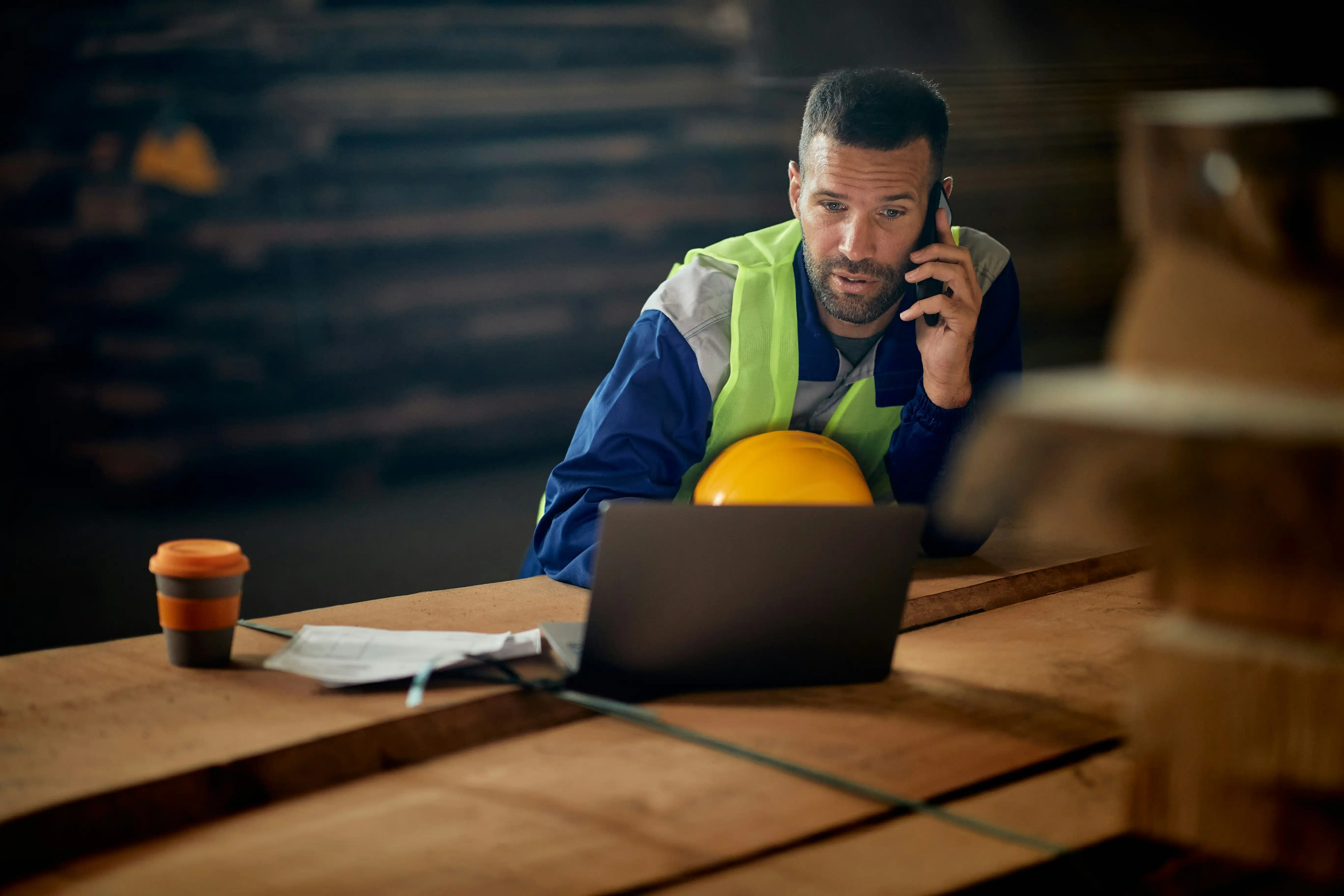 male warehouse worker wearing a green high vis jacket and blue overalls takes a phone call