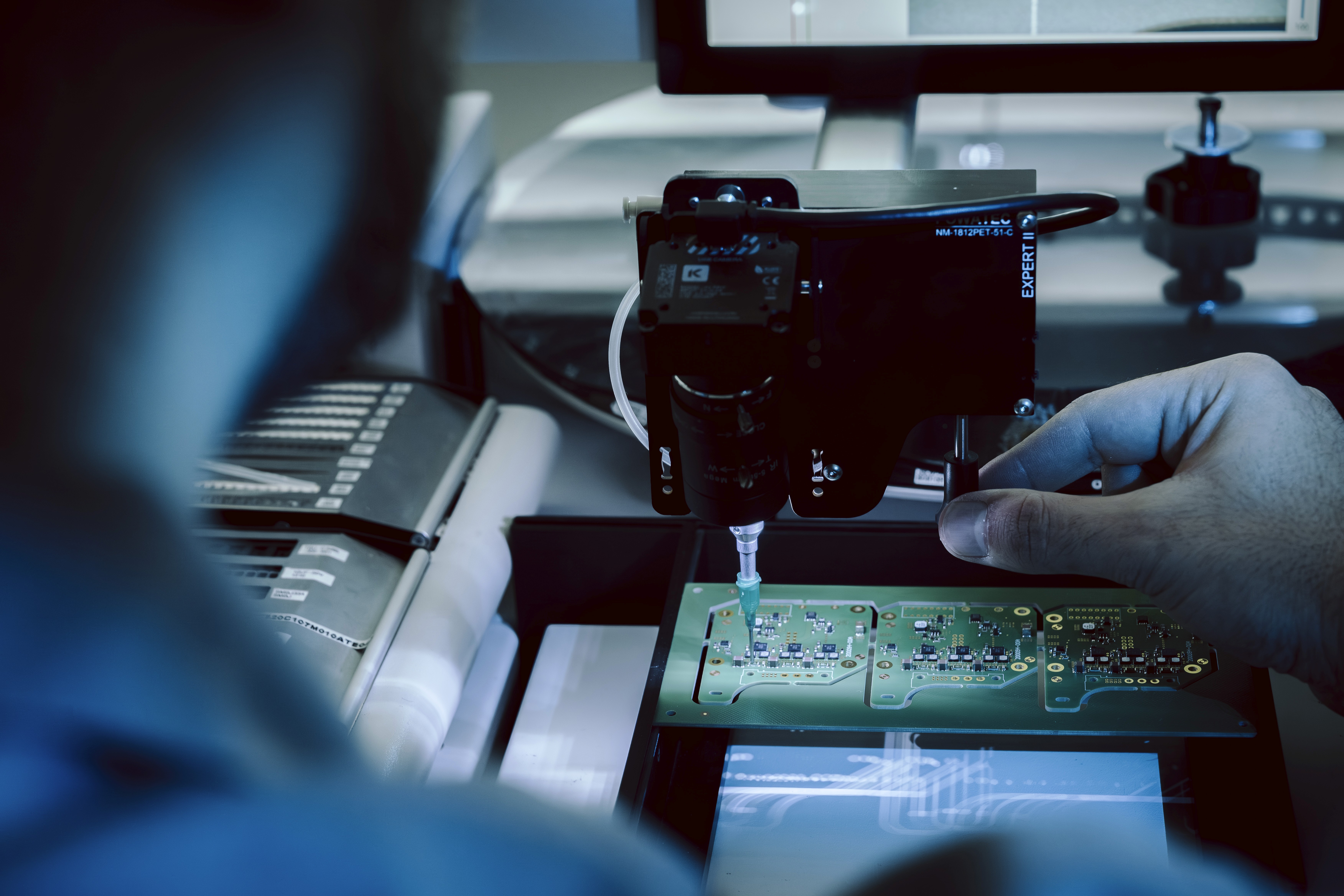 An over-the-shoulder view, past a person's blurred head and shoulder, focusing on their hand using thumb and index finger to precisely adjust a circuit board inside a complex machine.