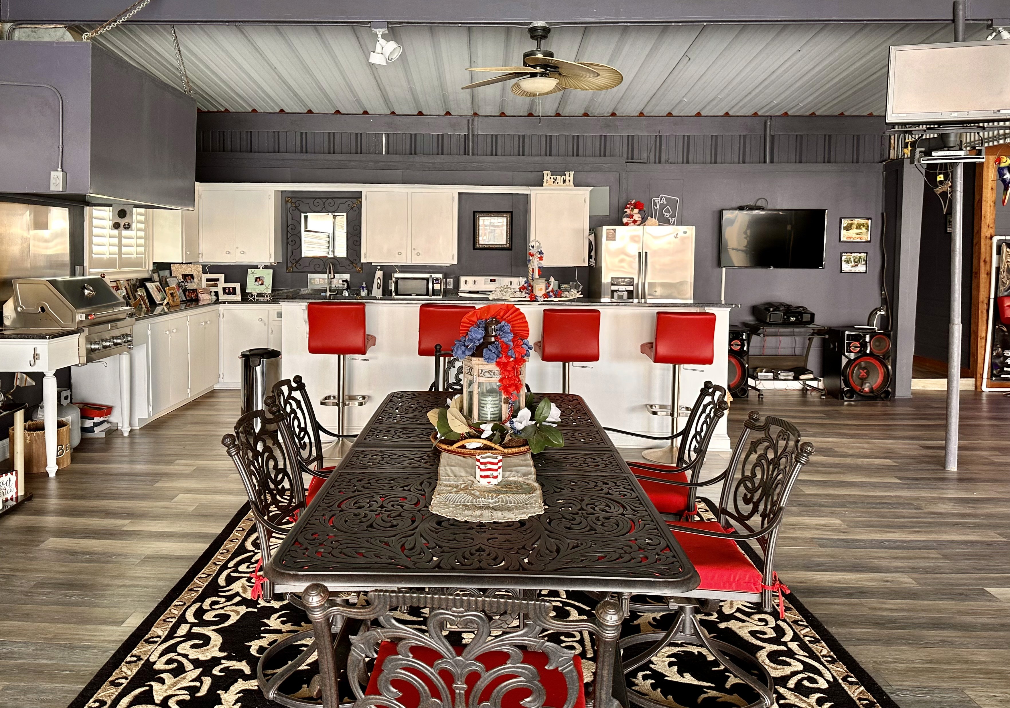 A modern kitchen and dining area features a decorative iron dining table with red cushioned chairs, surrounded by a patterned rug, with a backdrop of sleek gray and white cabinetry and stainless steel appliances.