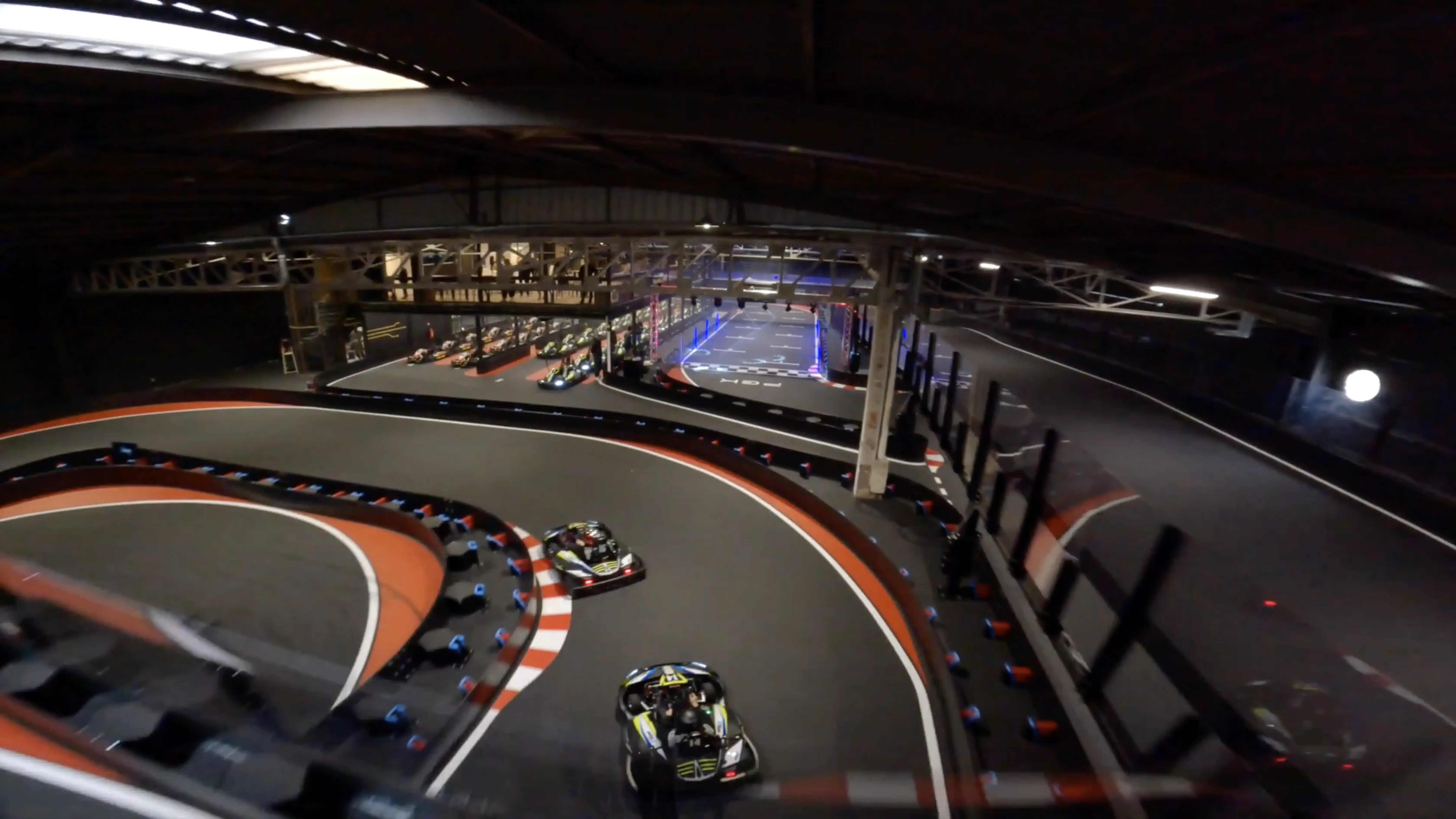 Aerial view of an indoor go-kart track with two karts racing around a curved section, featuring red and white striped barriers and illuminated overhead lights.