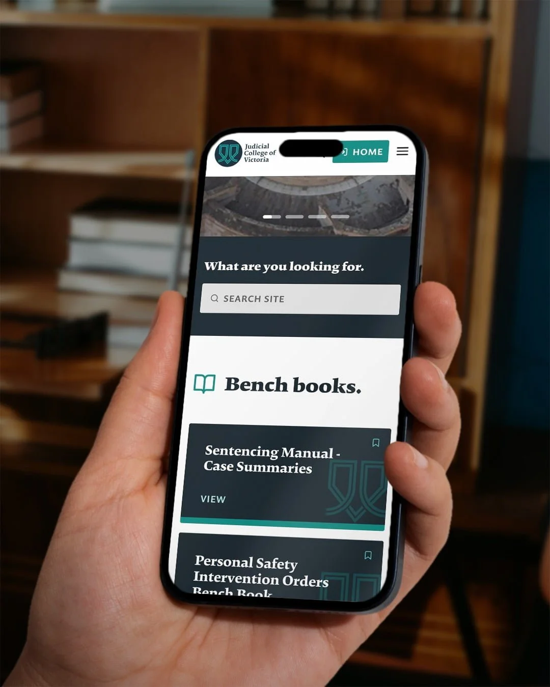 Hand holding a phone showing the Wisdom Shared search box and Bench books list for the Judicial College of Victoria