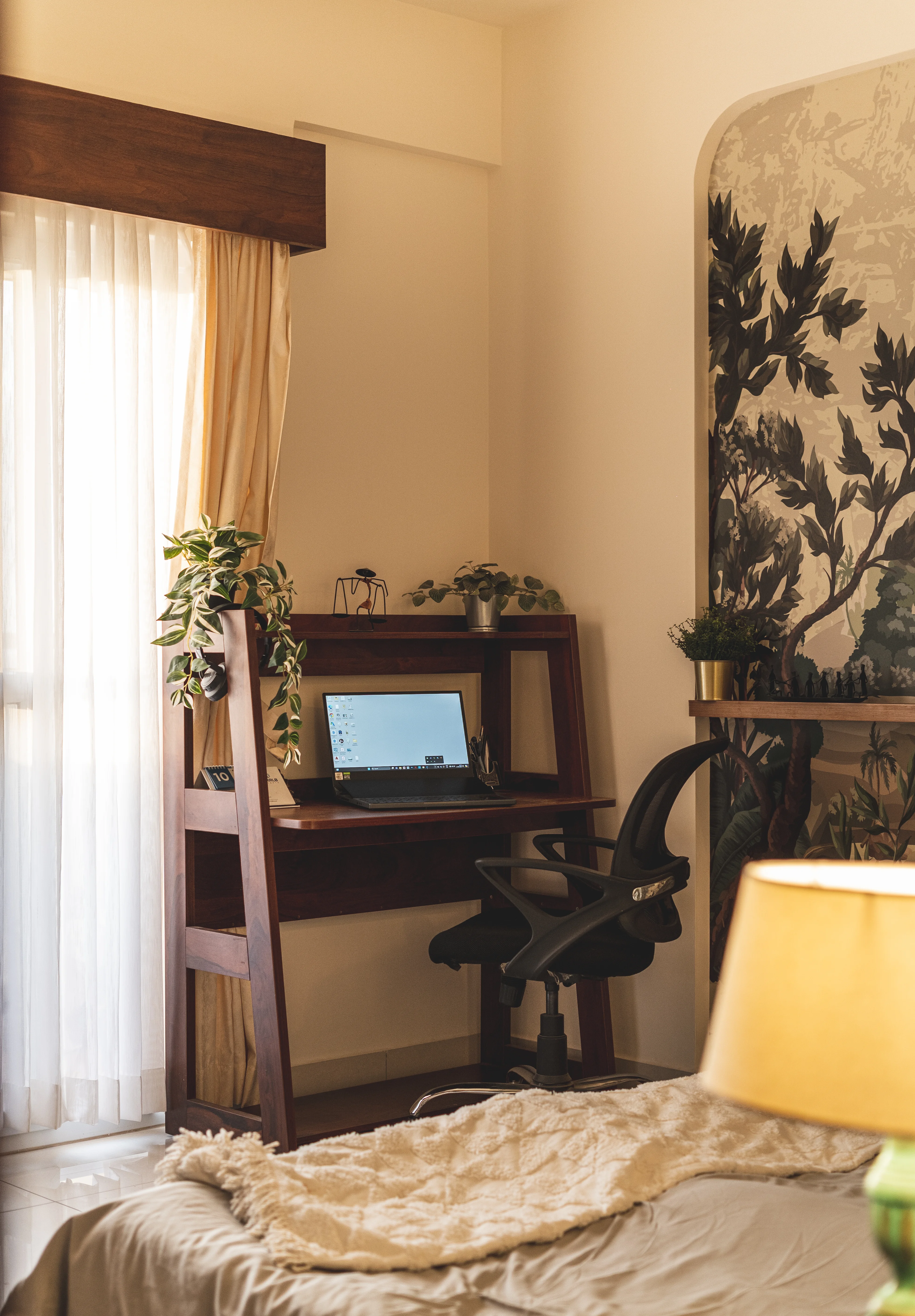A bedroom with a wooden desk positioned near a window. A laptop sits on the desk, and there are shelves for books and plants. A bed with white linens is visible in the background, along with a lamp and a tropical-themed wallpaper
