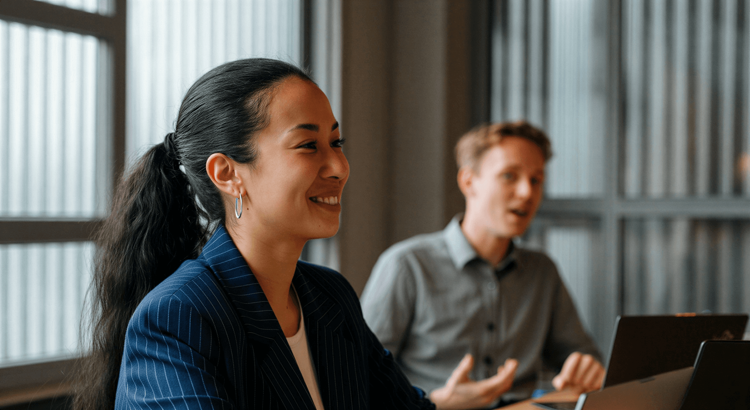Two professionals in a collaborative discussion, one smiling attentively while the other speaks animatedly in a bright office setting.