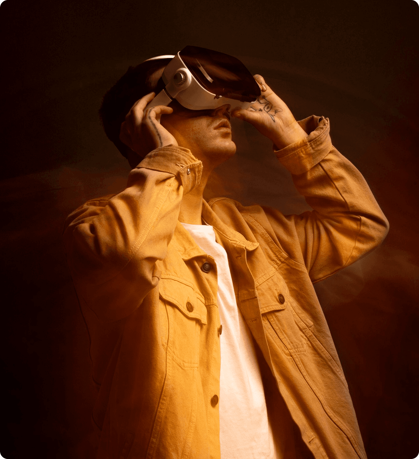 A cinematic, low-angle shot of a man wearing a white VR headset and a yellow denim jacket. He is looking upward with his hands adjusting the headset, set against a warm, smoky amber background with soft orange studio lighting.