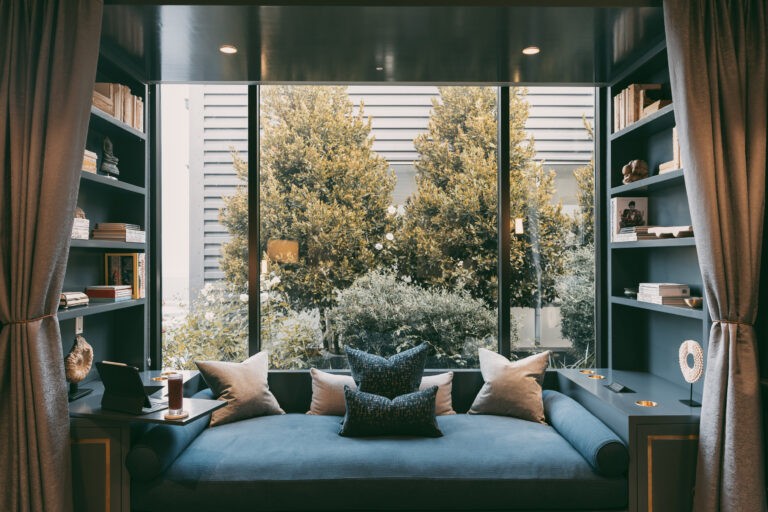 A cozy reading nook with a large window, plush blue seating, cushions, and bookshelves, offering a serene spot for relaxation and reflection.