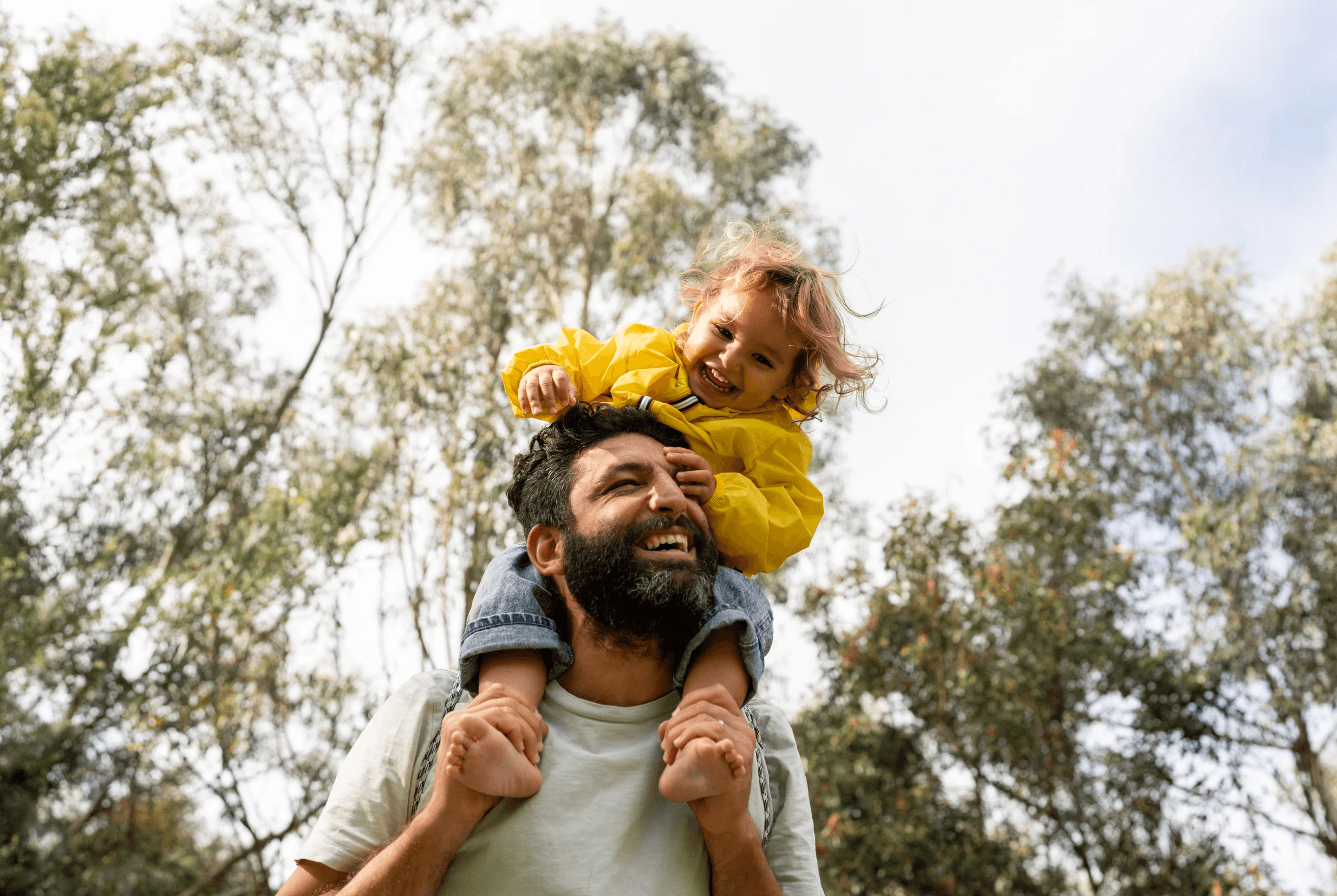 A dad smiles while holding his daughter on his shoulders in a wooded setting