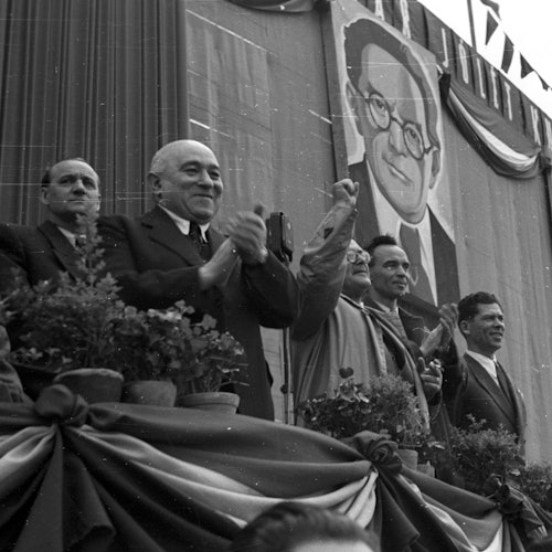 A group of men in suits standing on a decorated stage, clapping, with a large portrait of a bespectacled man in the background.