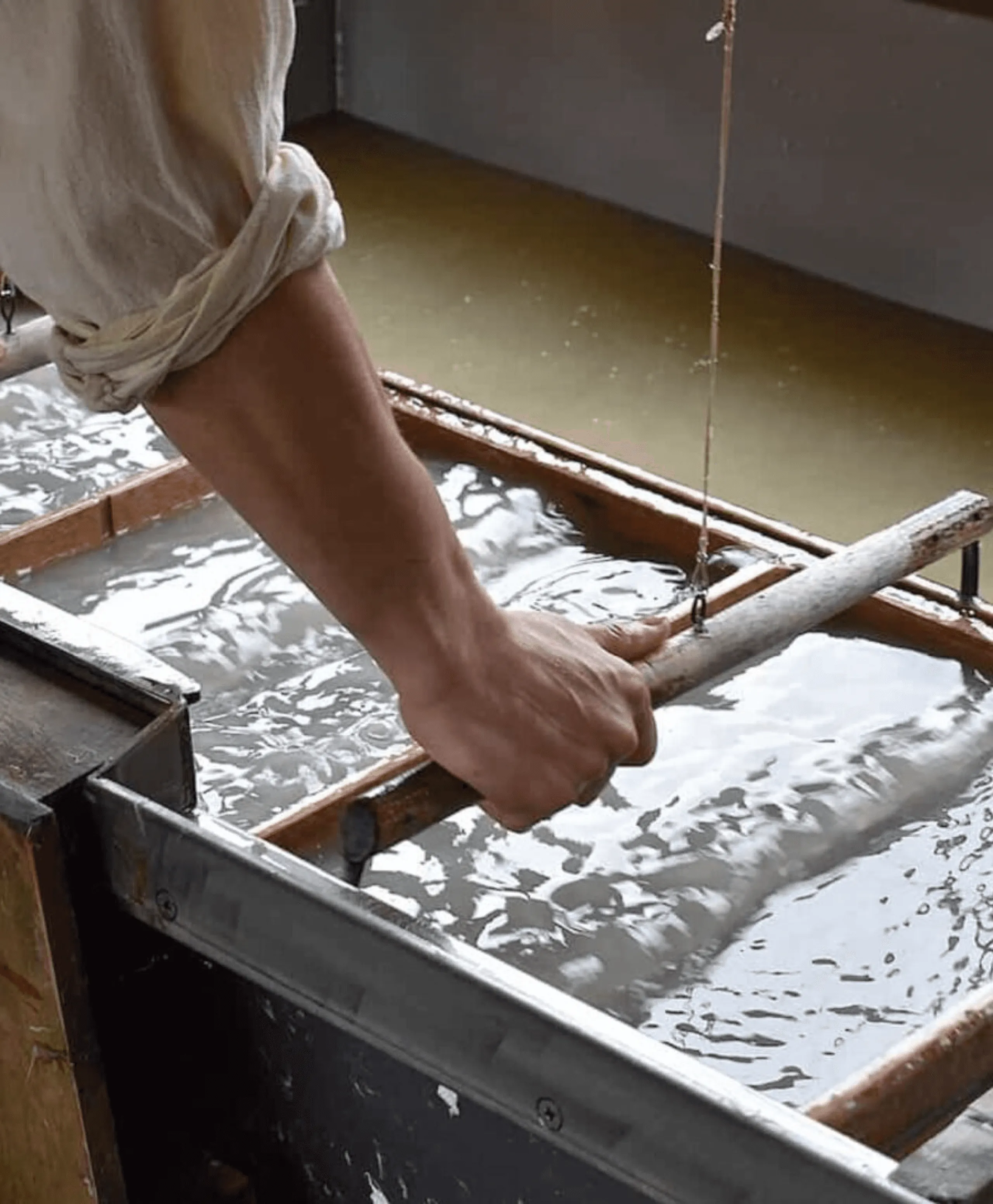 A close-up of a person's hands and arms making traditional handmade Japanese paper (washi). They are holding a large, rectangular wooden screen and dipping it into a vat of water and pulp to form a sheet.