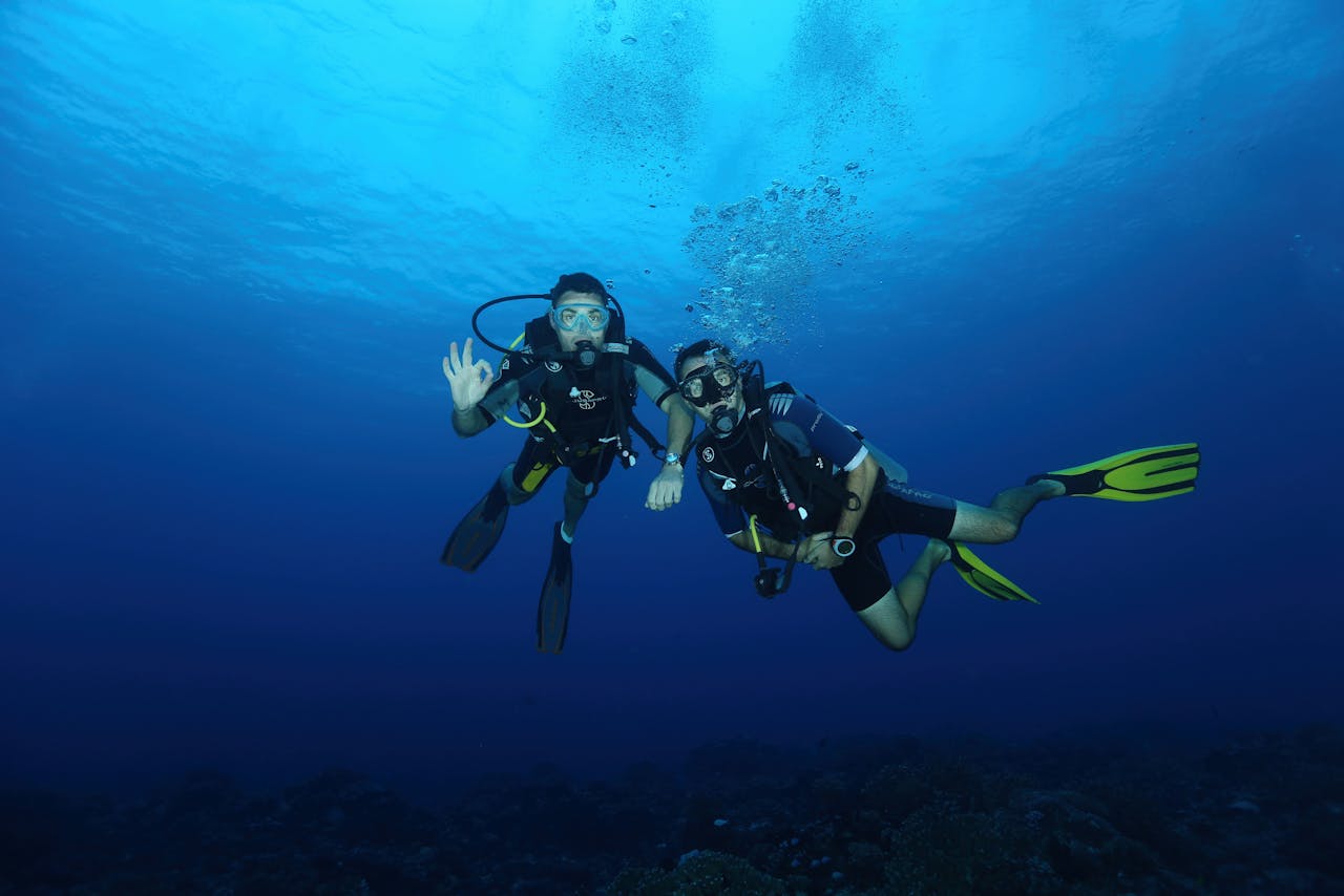 Two scuba divers swim together through deep waters, wearing full diving gear.