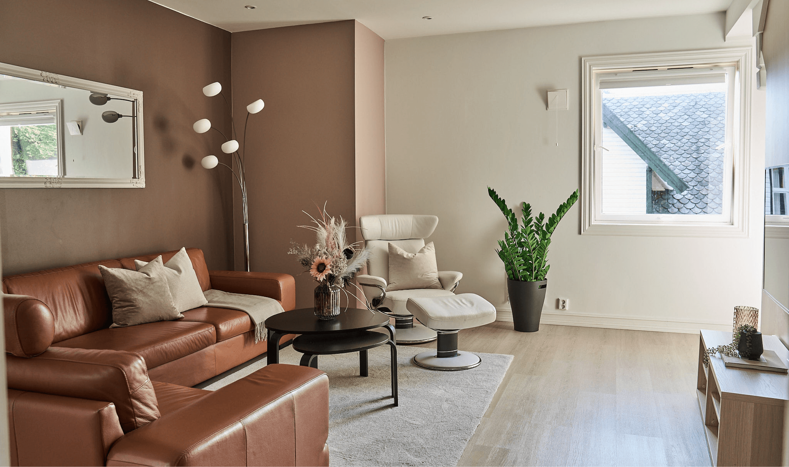 An image from one of the apartments. Showing a livingroom with a brown leather sofa, a black coffee table and a white lounge chair. A tall green plant in the center and a tall lamp in the corner. A window to the right. There is also a carpet on the floor and one of the walls are painted brown. 