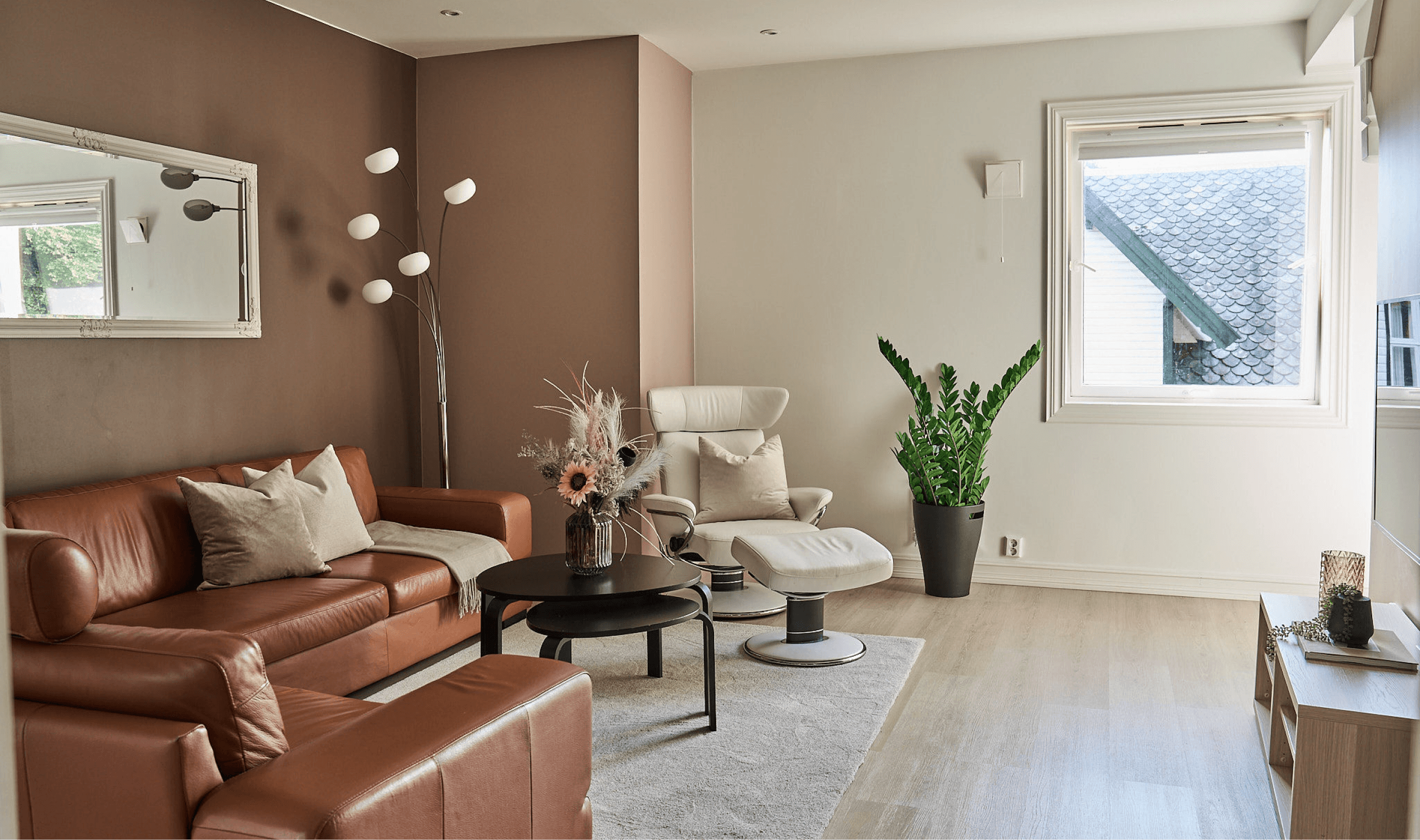 An image from one of the apartments. Showing a livingroom with a brown leather sofa, a black coffee table and a white lounge chair. A tall green plant in the center and a tall lamp in the corner. A window to the right. There is also a carpet on the floor and one of the walls are painted brown.