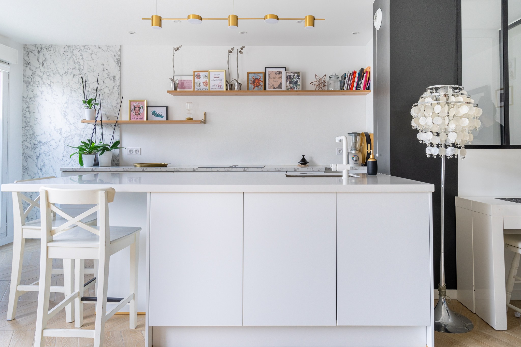 Cuisine moderne avec un îlot blanc, des sols en bois clair, un tabouret, des plantes, des œuvres d'art murales sur des étagères flottantes et une lampe décorative avec des coquillages suspendus. Des touches de marbre et un décor minimaliste complètent l'espace.
