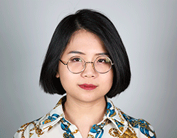 The image shows a person with short, dark hair and glasses. The individual is wearing a white shirt with a blue and gold pattern. The background is a plain light gray or off-white color. The person's expression is neutral or slightly serious. They are looking directly at the camera. The person has dark eyebrows and dark eyelashes. The glasses have thin metal frames.