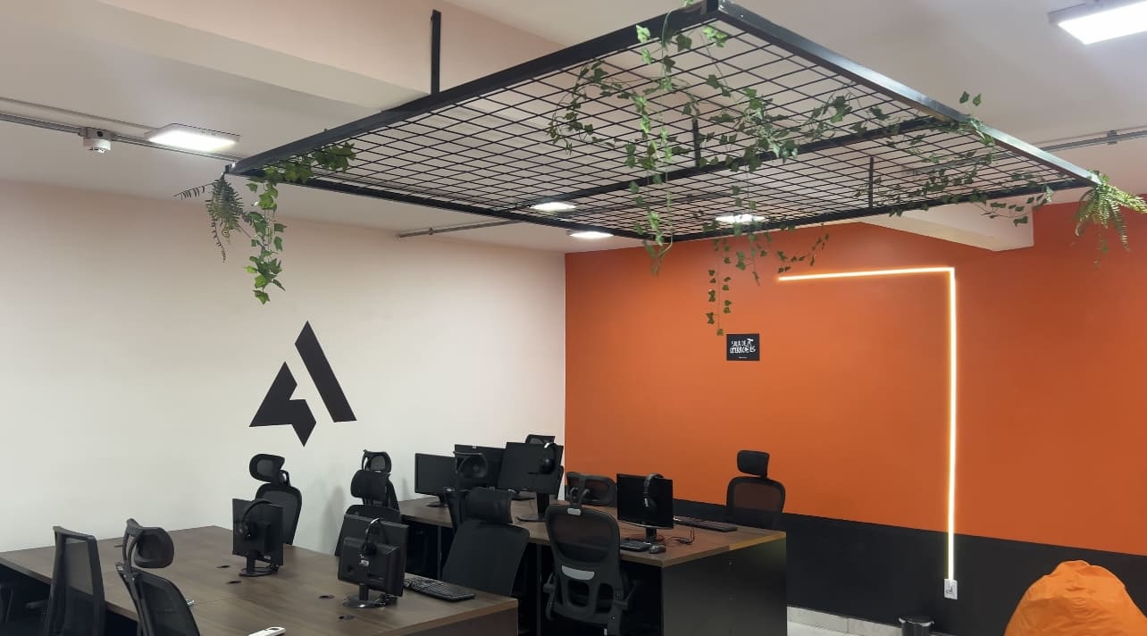 Escritório moderno com estações de trabalho, computadores, cadeiras ergonômicas, parede laranja e plantas suspensas, transmitindo inovação e alta performance.