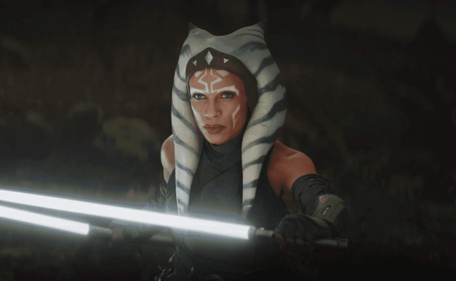 Ahsoka with white and gray head-tails holds a glowing saber, looking determined in a dark setting.