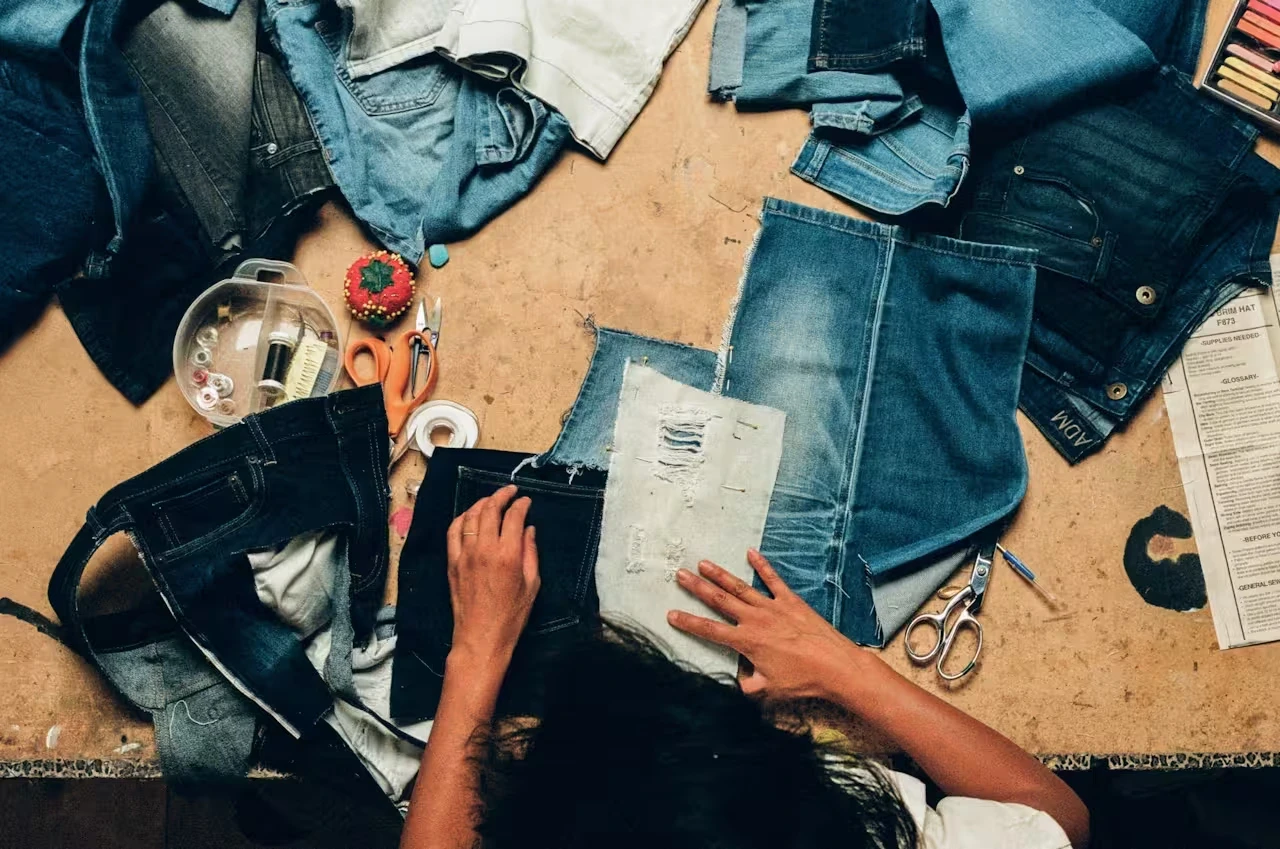 Overhead view of a person working on denim fabric pieces surrounded by scissors, a pincushion, and sewing notions.