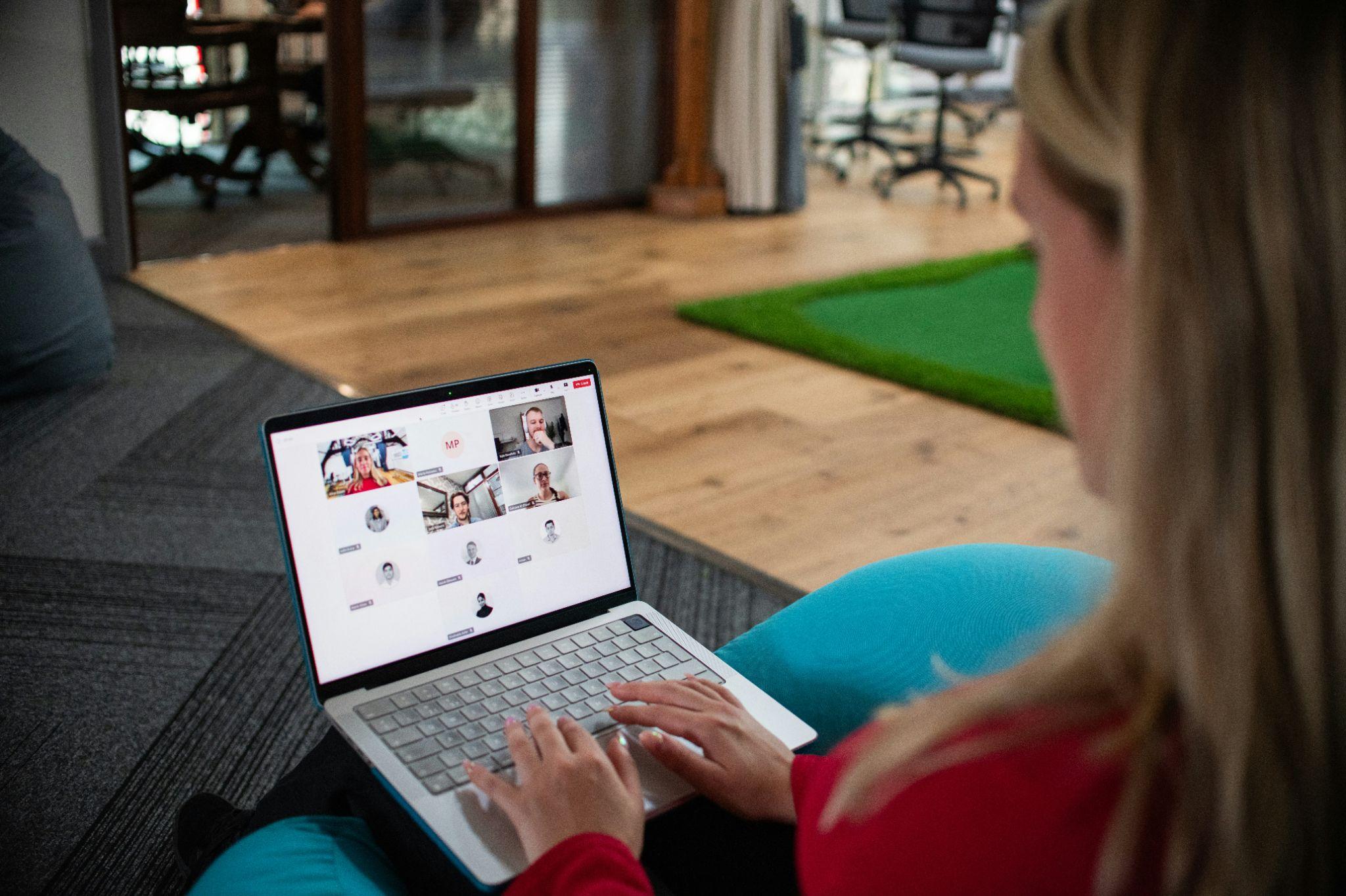 Persona participando en una videollamada grupal desde una laptop, en un espacio de oficina con suelo de madera y mobiliario moderno.