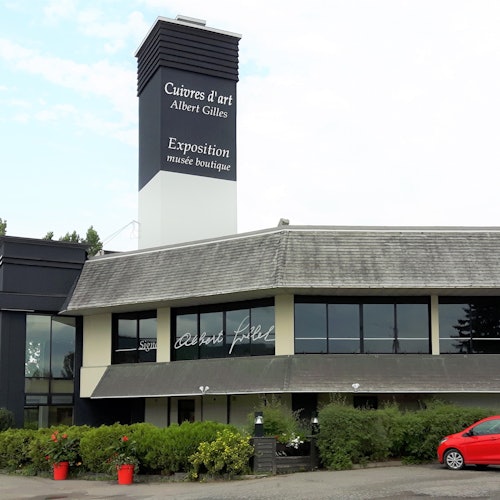 Một tòa nhà với biển hiệu cao có dòng chữ "Cuivres d'art Albert Gilles," có cửa hàng bảo tàng. Có các bụi cây và một chiếc ô tô màu đỏ ở phía trước.