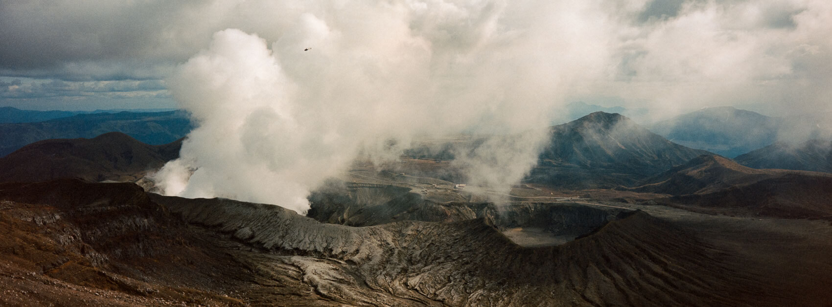 Japan hiking at Mount Aso: two volcanic craters creating clouds over the unworldly landscape with a tiny helicopter flying near it showing scale, captured on Kodak Portra film in Kyushu's otherworldly terrain.