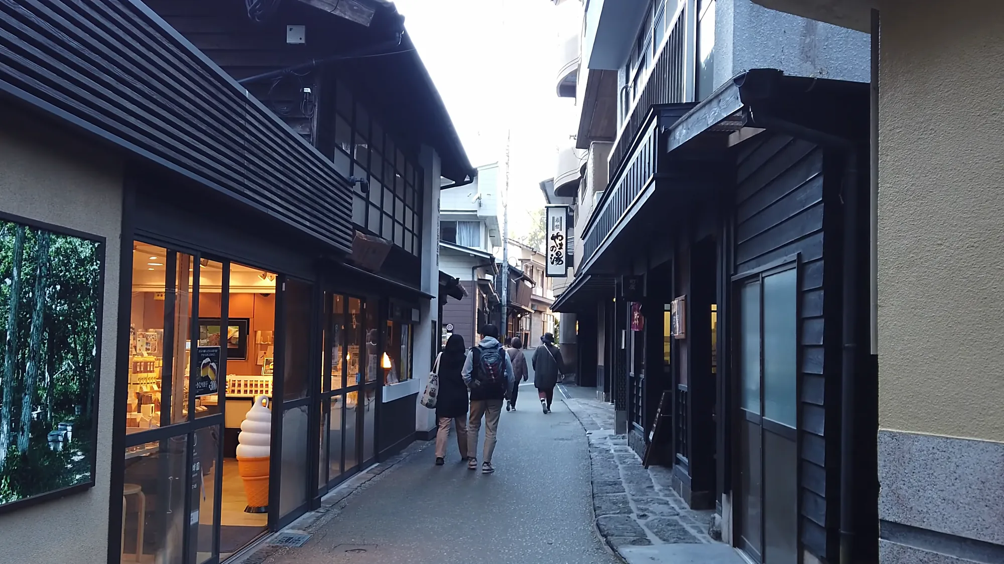 Walkers on the evening lane at Kurokawa Onsen past lit shopfronts under a grey sky
