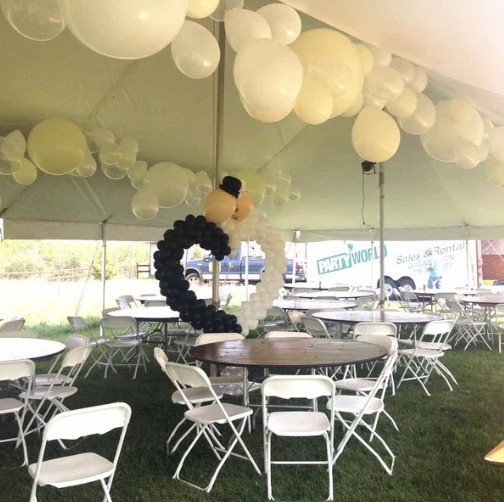 A large outdoor party tent is decorated with clusters of white and yellow balloons, surrounding rows of white folding chairs and tables set on a grassy field.