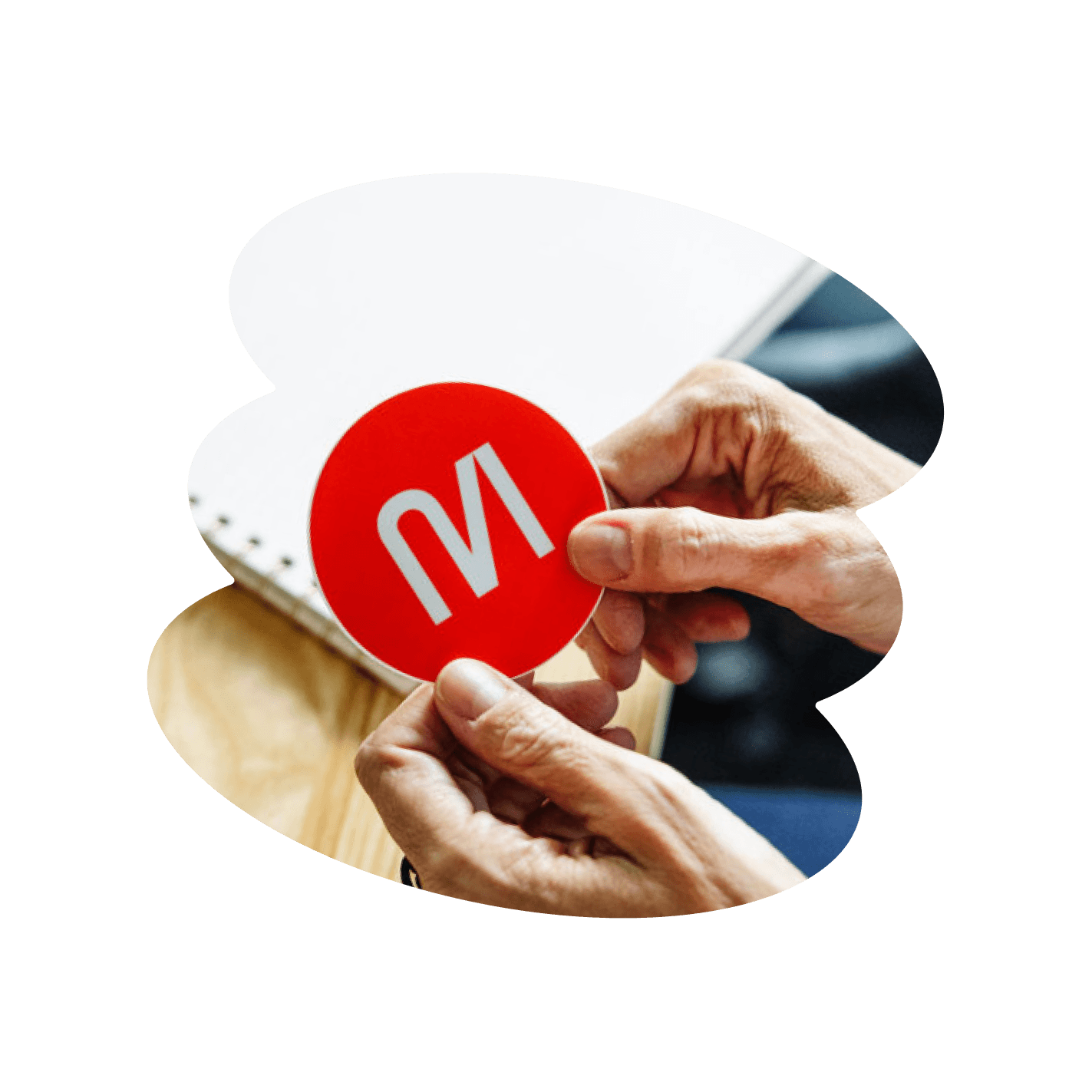 Hands holding a red circular logo with a white “M” over a wooden table and notebook background.