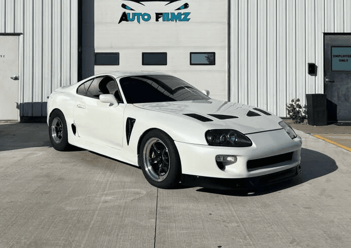 A white Toyota Supra sports car with black wheels and a sleek, modified design is parked in front of a white industrial building.