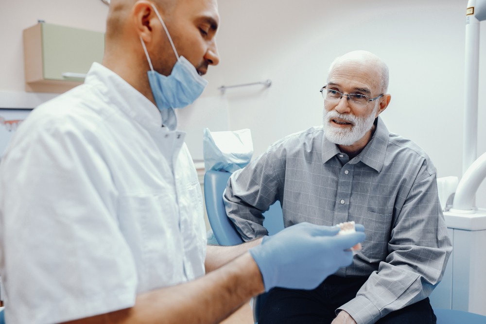 A dentist wearing a mask and gloves explains a dental model to an older man seated in a dental chair.