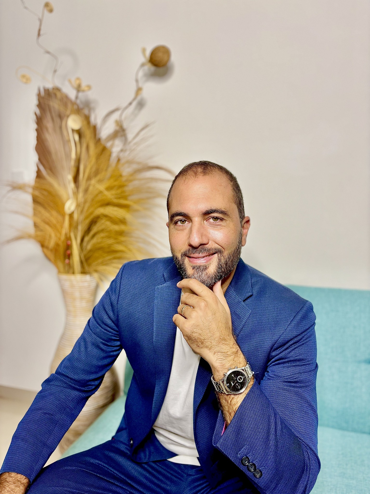 Portrait of Younis Hijazi, PhD, Co-founder of Invest Indo Group, sitting comfortably in a blue suit, with a background featuring tropical décor.