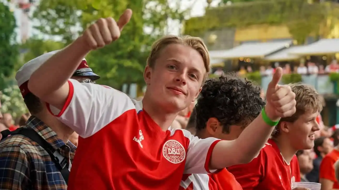 A young Danish fan in a red jersey joyfully gives a thumbs-up during a UEFA European Championship 2024 match, surrounded by other cheering spectators.