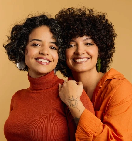 Duas mulheres utilizando roupas nos tons de laranja, posando para uma foto institucional.