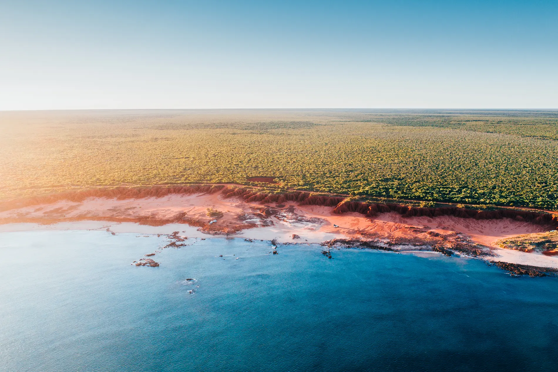 Aerial at low sun over a scrub-covered coast, pale sand edging shallow blue water