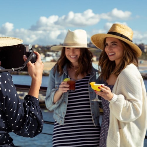 Two women wearing hats smile at the camera, holding drinks, with a person photographing them by a seaside backdrop.